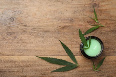 Photo of Jar of hemp cosmetics with green leaves on wooden table, flat lay. Space for text