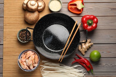 Black wok and chopsticks surrounded by products on color wooden table, flat lay