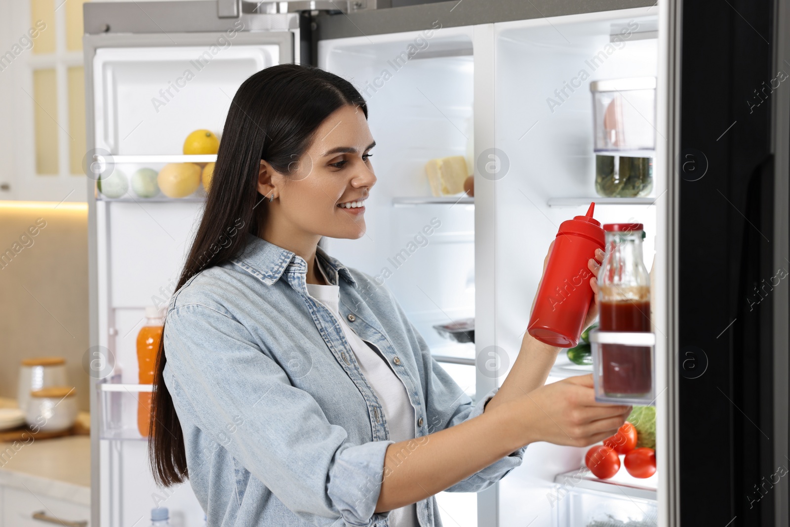 Photo of Young woman taking bottle of ketchup out of refrigerator in kitchen