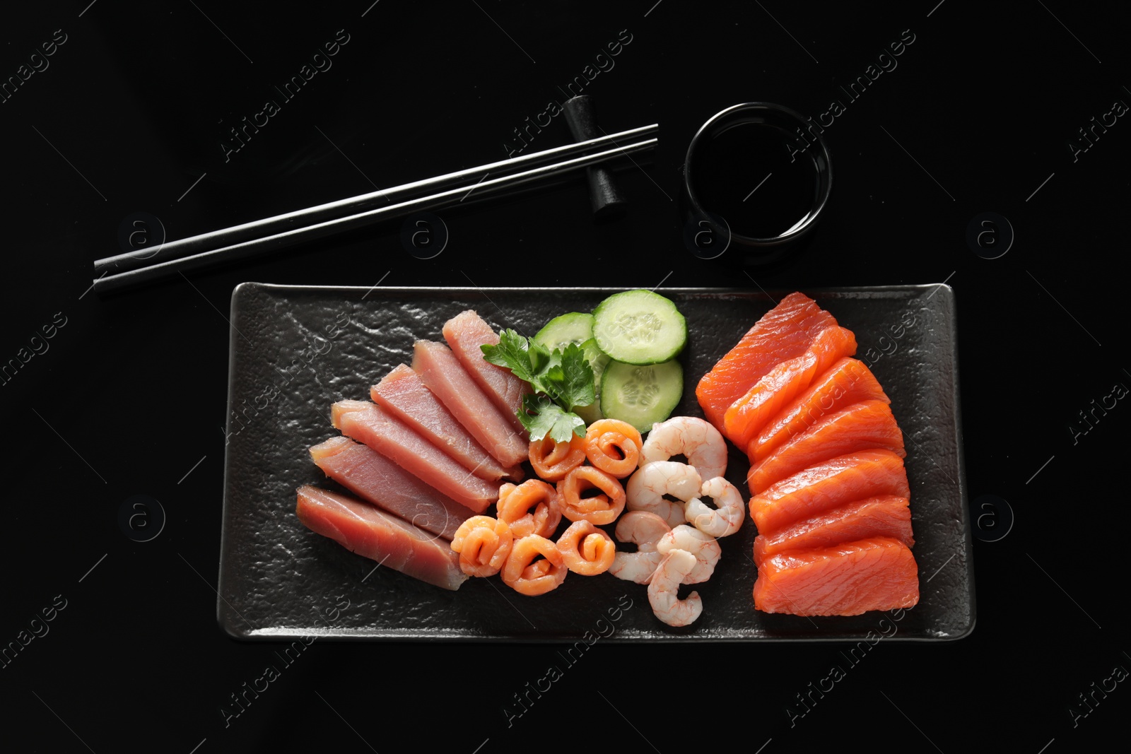 Photo of Delicious sashimi set of salmon, tuna and shrimps served with cucumbers, parsley and soy sauce on black background, flat lay