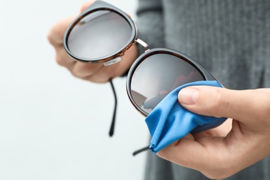 Woman wiping sunglasses with microfiber cleaning cloth on light background, closeup