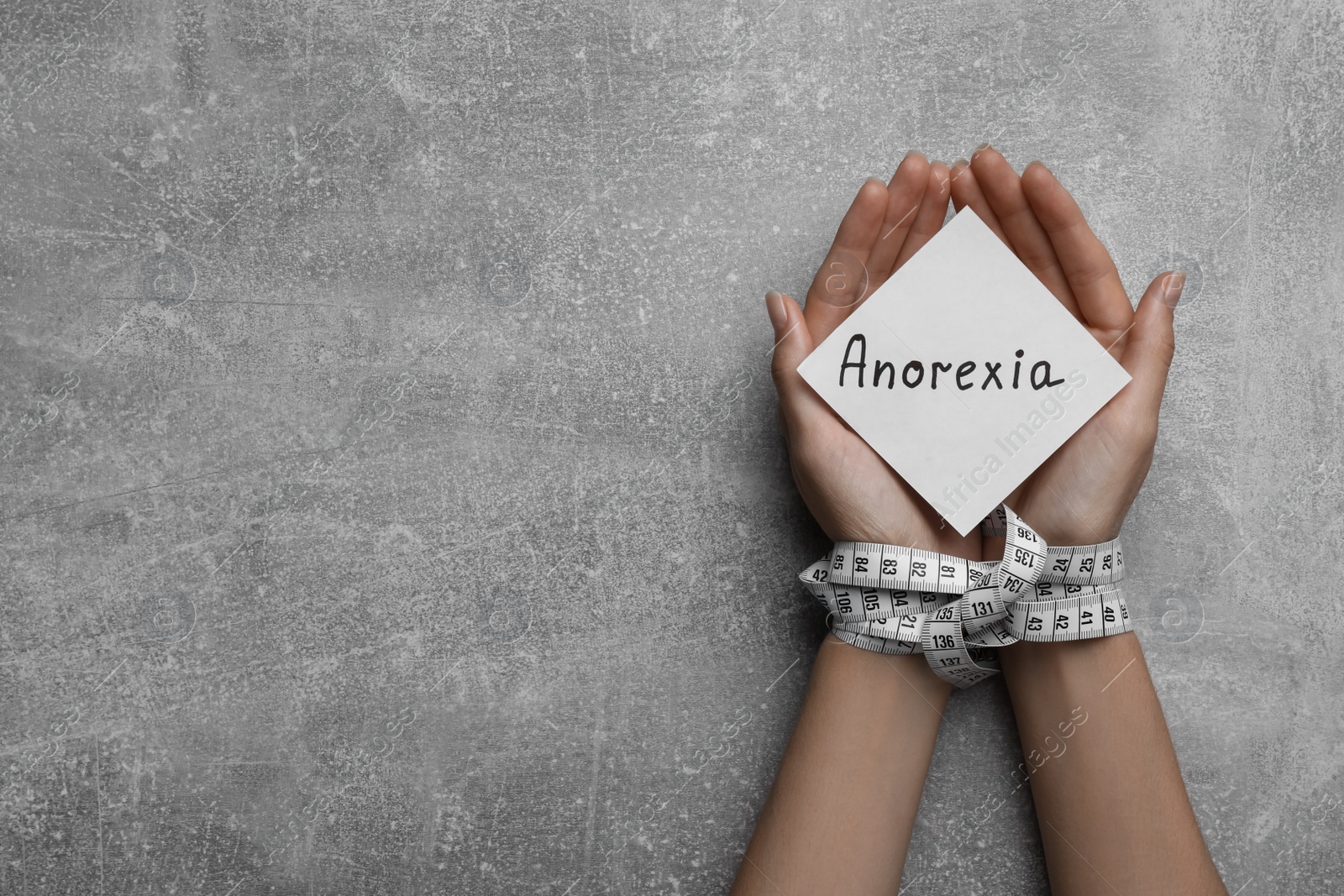 Photo of Woman holding paper note with word Anorexia in hands tied with measuring tape, top view. Space for text