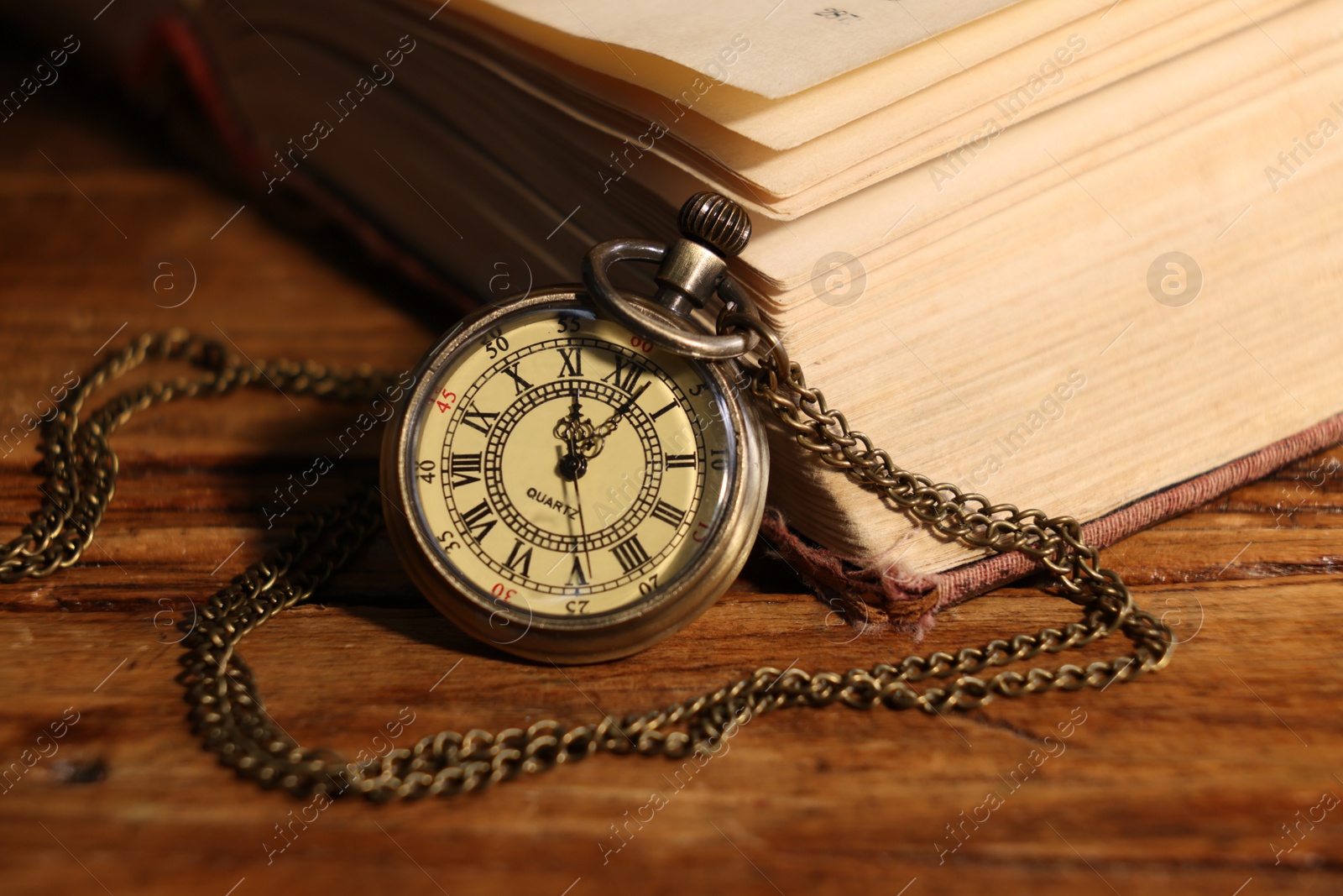 Photo of Pocket clock with chain and book on wooden table, closeup