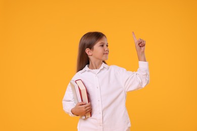 Photo of Cute schoolgirl with books pointing at something on orange background