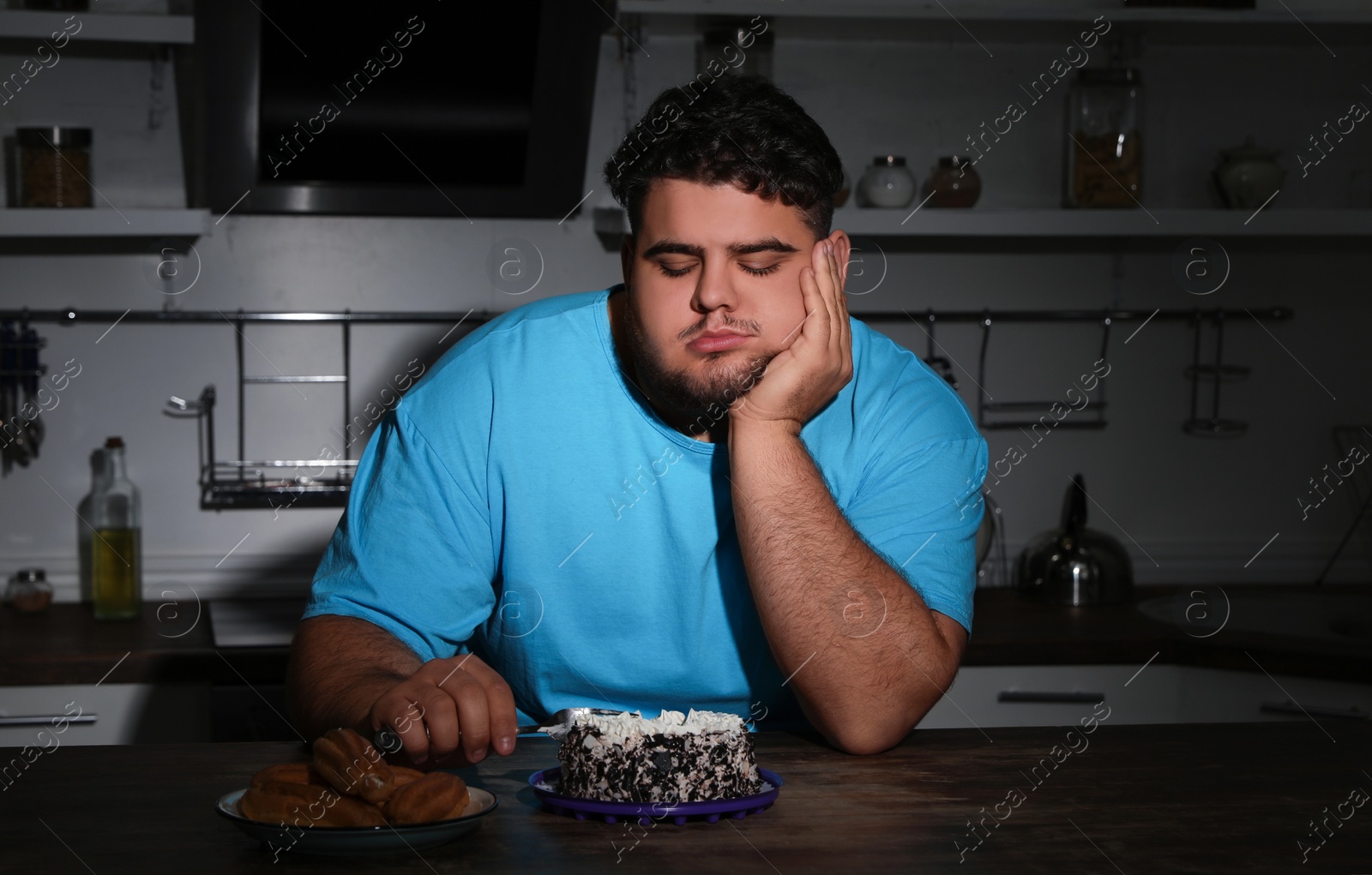 Photo of Depressed overweight man eating cake in kitchen at night