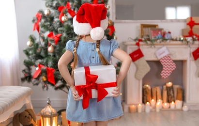 Cute little child in Santa hat hiding Christmas gift box behind back at home