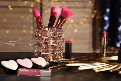 Photo of Different beautiful lipsticks, brushes and highlighters on table against blurred lights