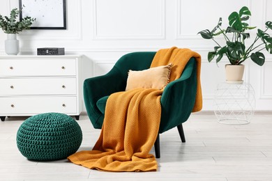 Comfortable armchair with blanket, ottoman, houseplant and chest of drawers in room