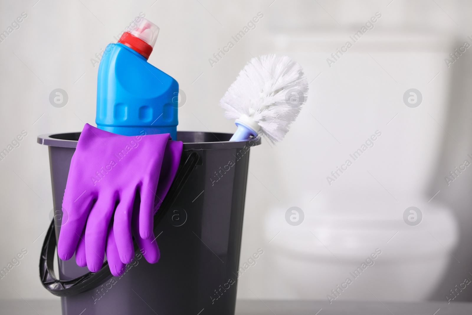 Photo of Bucket with bottle, toilet brush and gloves on table indoors, space for text. Cleaning supplies