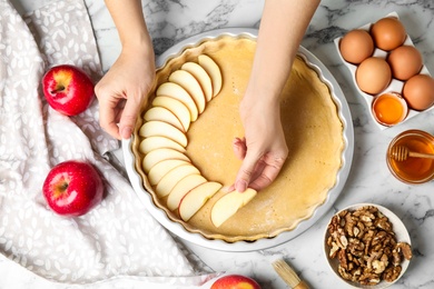 Photo of Woman putting apple slice into baking dish with dough to make traditional English pie at white marble table, top view