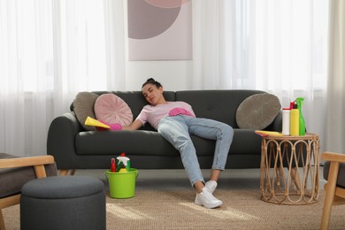 Photo of Tired young woman sleeping on sofa and cleaning supplies in living room
