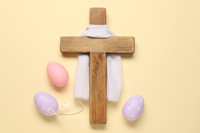 Photo of Wooden cross, white cloth and painted Easter eggs on beige background, flat lay