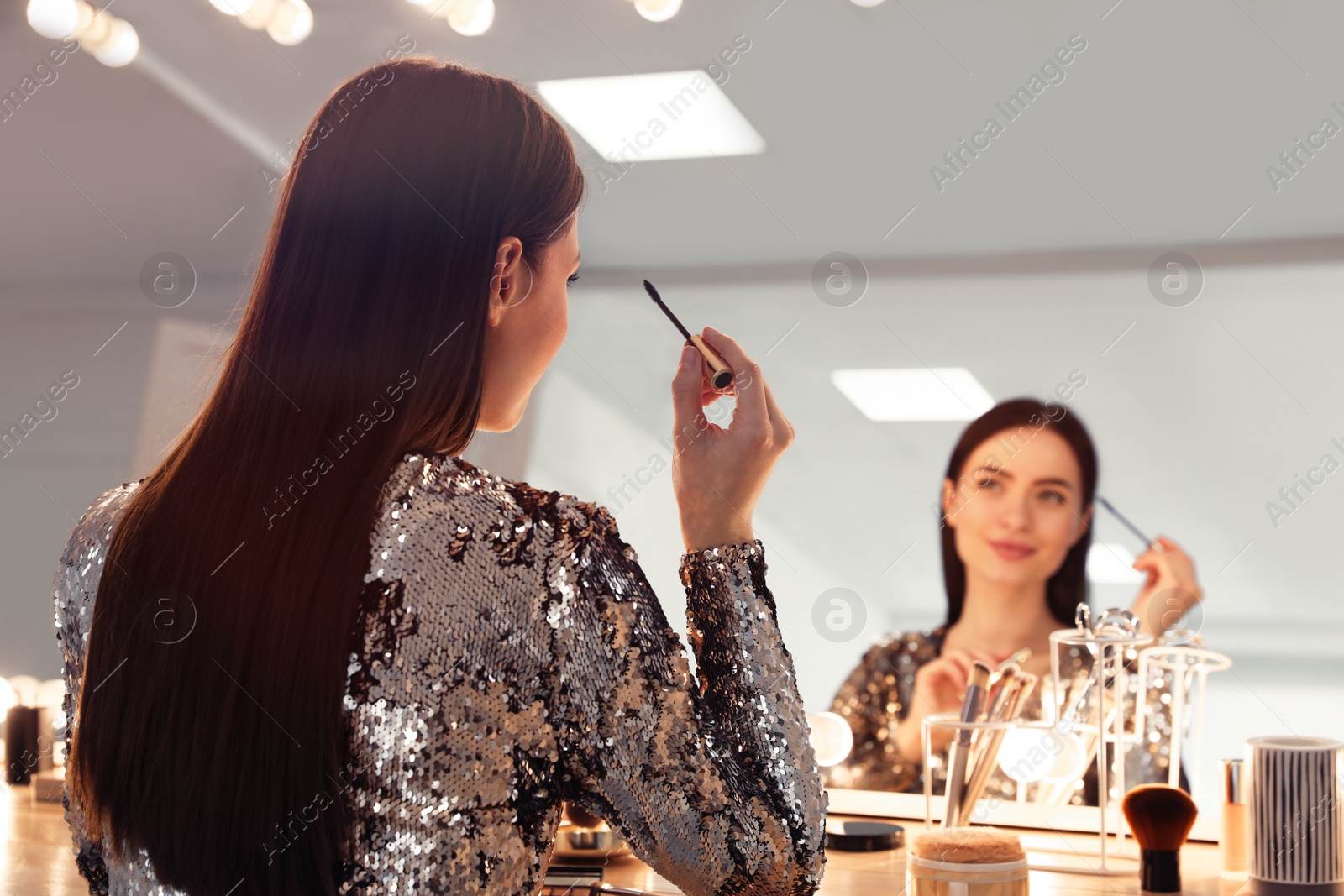 Photo of Young woman applying make up near illuminated mirror indoors