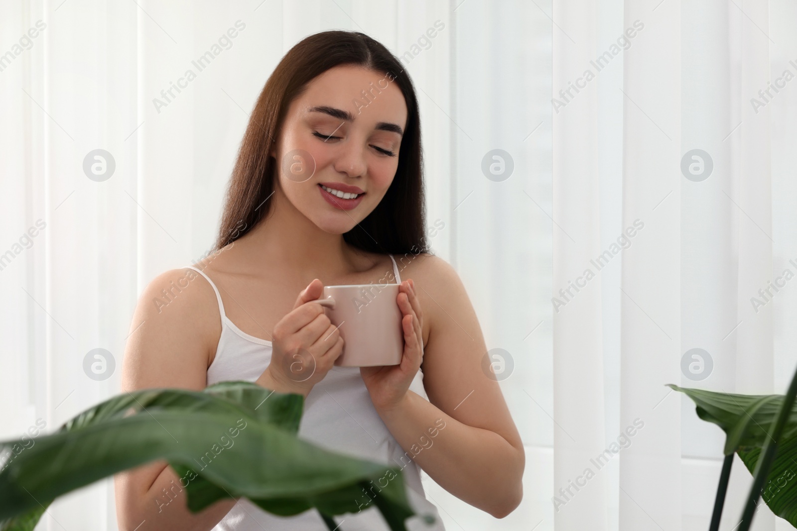 Photo of Happy woman with cup of drink near window indoors, space for text. Lazy morning