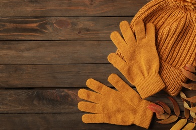 Stylish orange woolen gloves, hat and dry leaves on wooden table, flat lay. Space for text