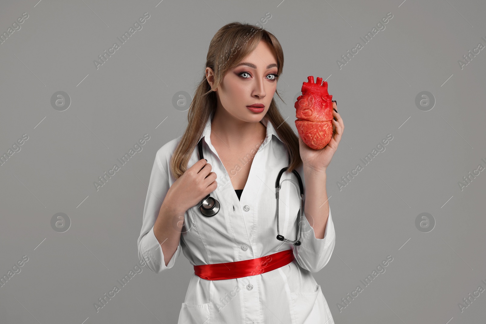 Photo of Woman in scary nurse costume with heart model on light grey background. Halloween celebration