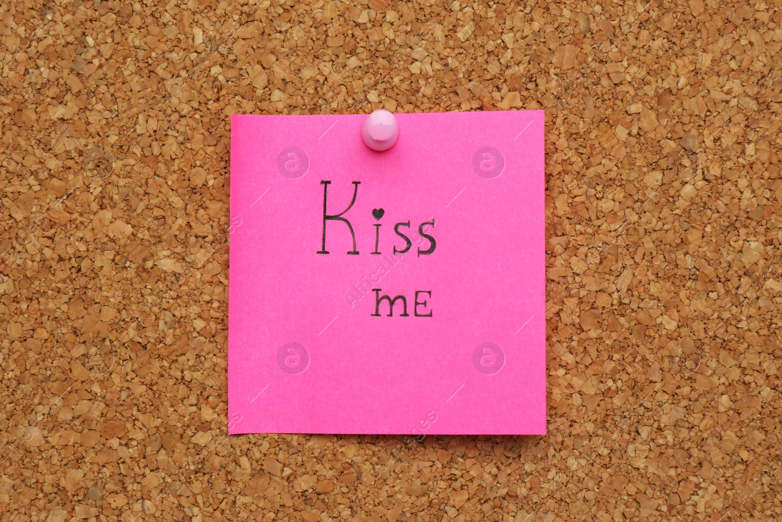 Photo of Pink paper note with phrase Kiss Me pinned to cork board