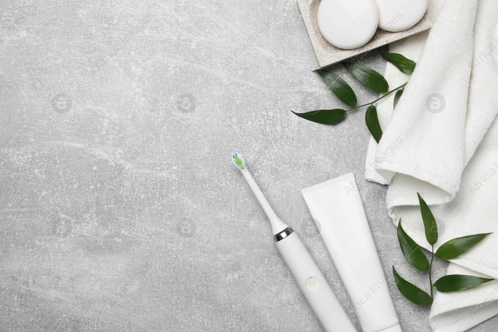 Photo of Electric toothbrush, tube of paste, towel and leaves on grey textured table, flat lay. Space for text