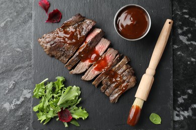 Photo of Pieces of delicious roasted beef meat with sauce and greens on black table, top view