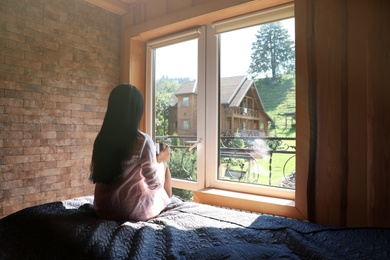 Photo of Young woman sitting on bed and enjoying view from window. Peaceful morning