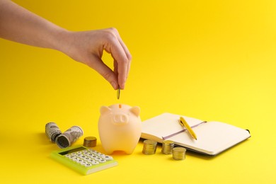 Financial savings. Woman putting coin into piggy bank on yellow background, closeup