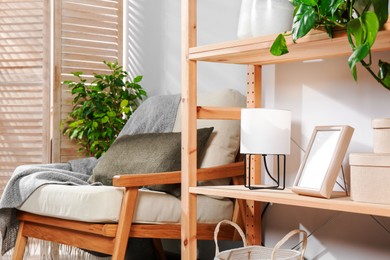 Photo of Wooden shelving unit with home decor, folding screen and armchair near light wall in room. Interior design