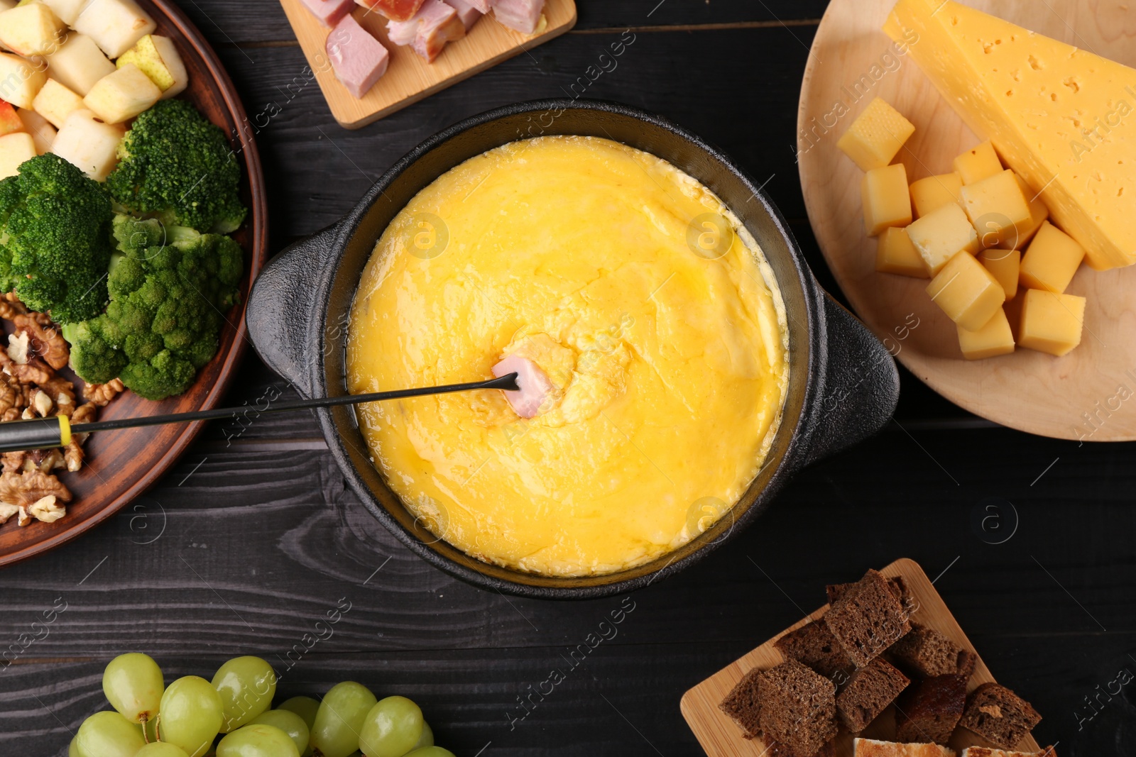 Photo of Fondue pot with melted cheese, fork and different products on black wooden table, flat lay