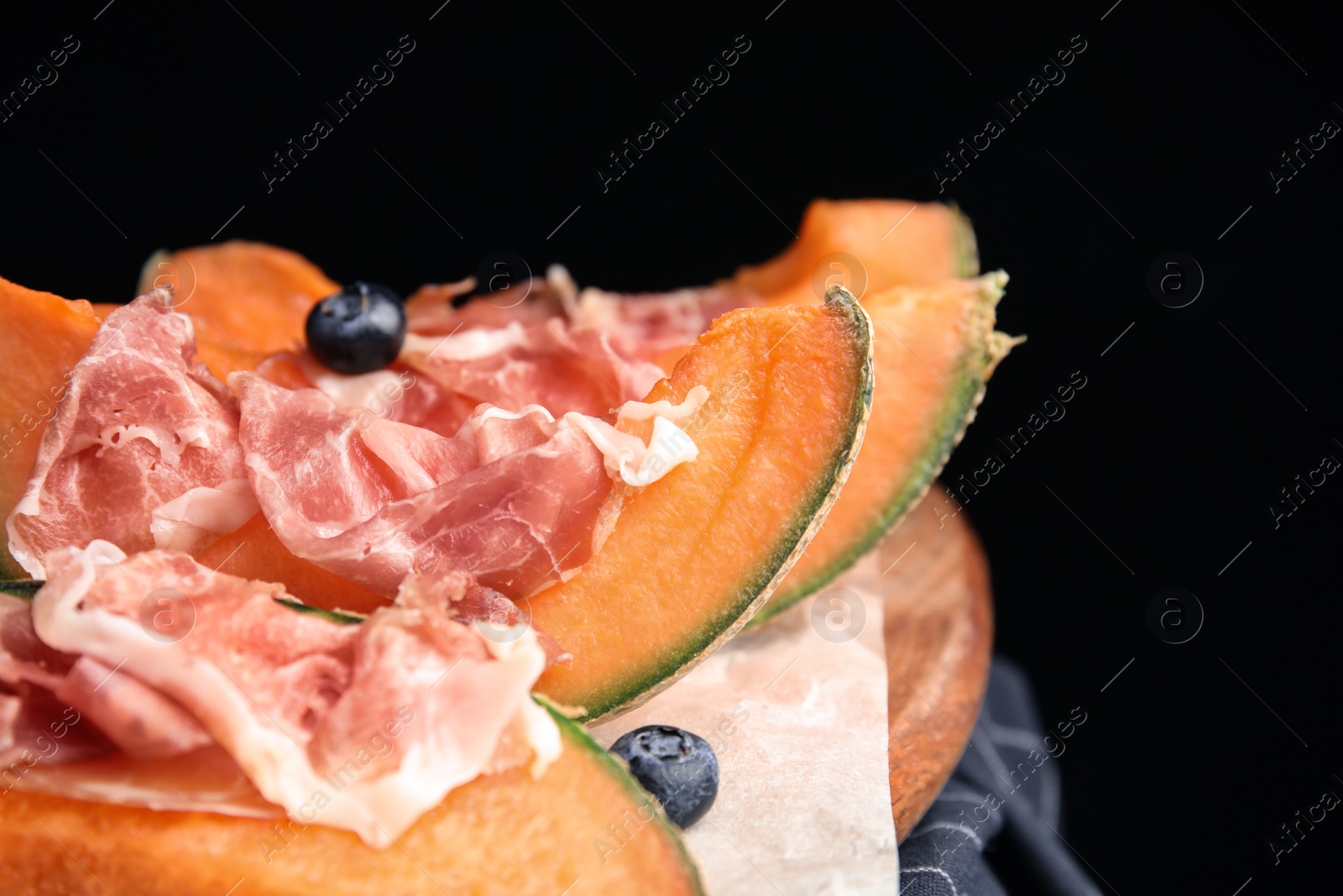 Photo of Wooden board with melon, prosciutto and blueberries on table against black background