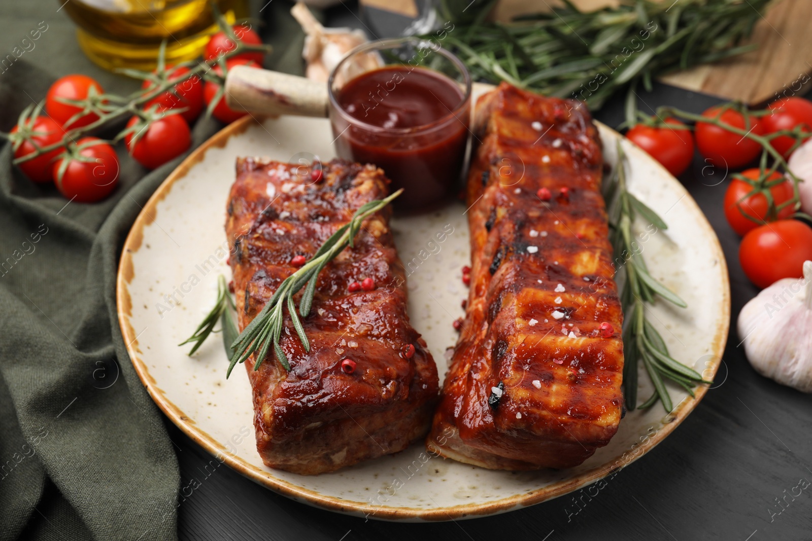 Photo of Tasty roasted pork ribs served with sauce, tomatoes and other products on grey wooden table, closeup