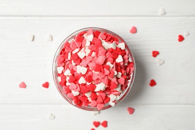 Photo of Bright heart shaped sprinkles in glass bowl on white wooden table, flat lay