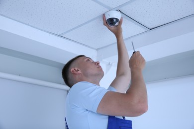 Technician with screwdriver installing CCTV camera on ceiling indoors