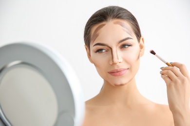 Photo of Young woman applying contouring lines on her face against light background. Professional makeup products