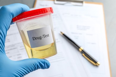 Photo of Drug test. Laboratory worker holding container with urine sample over medical forms on table, closeup