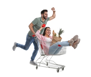 Young man giving his girlfriend ride in shopping cart with groceries on white background