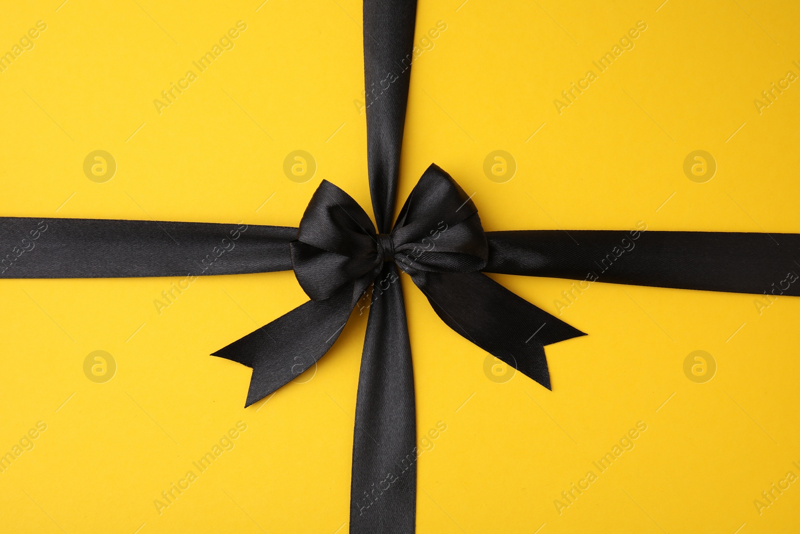 Photo of Black satin ribbon with bow on yellow background, top view