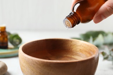 Photo of Woman dripping eucalyptus essential oil from bottle into bowl on blurred background, closeup