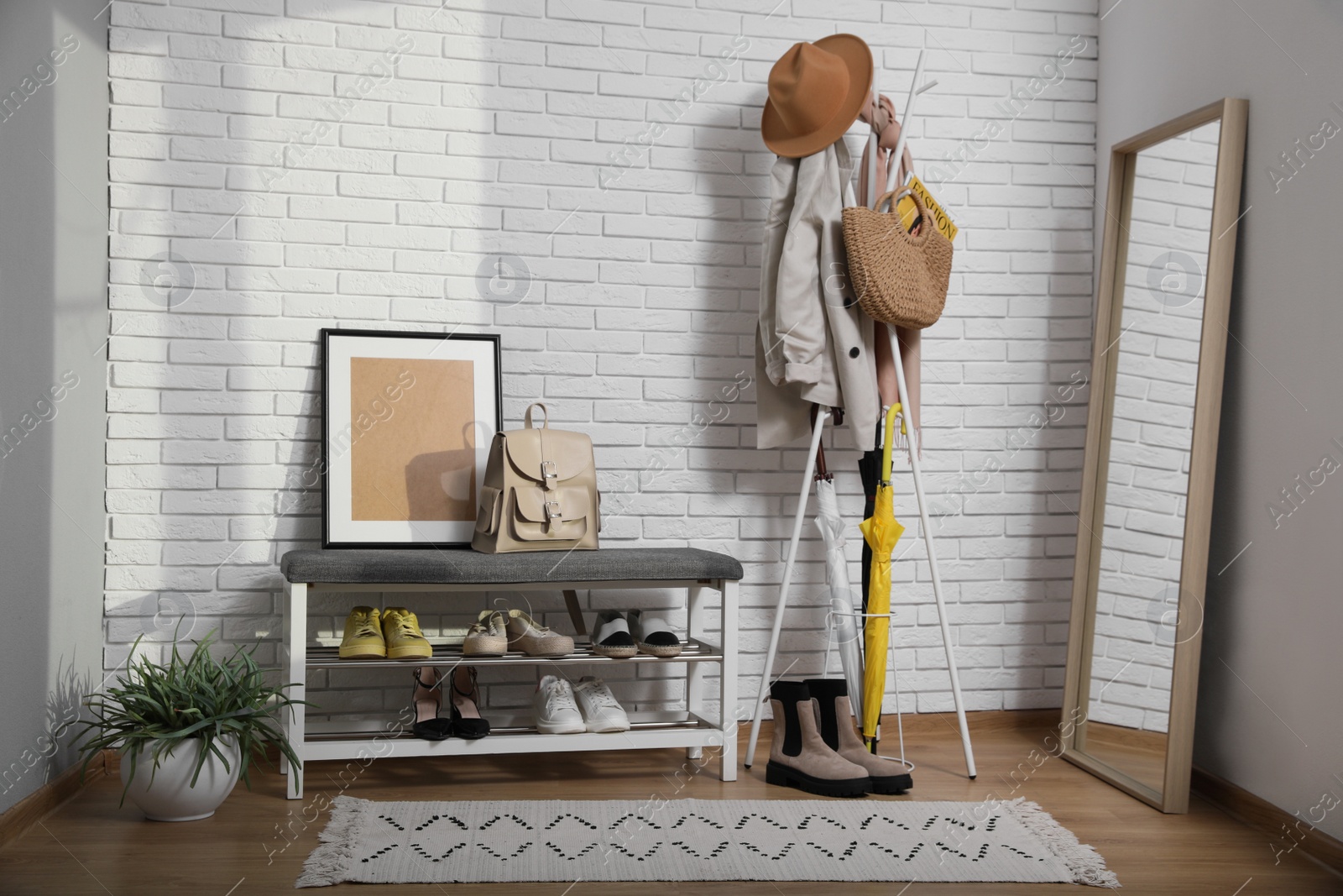 Photo of Stylish hallway interior with coat rack, shoe storage bench and mirror near white brick wall