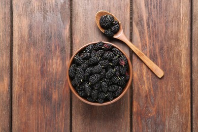 Bowl and spoon with delicious ripe black mulberries on wooden table, flat lay