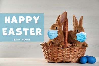 Image of Text Happy Easter Stay Home and cute bunnies in protective masks on white table. Holiday during Covid-19 pandemic