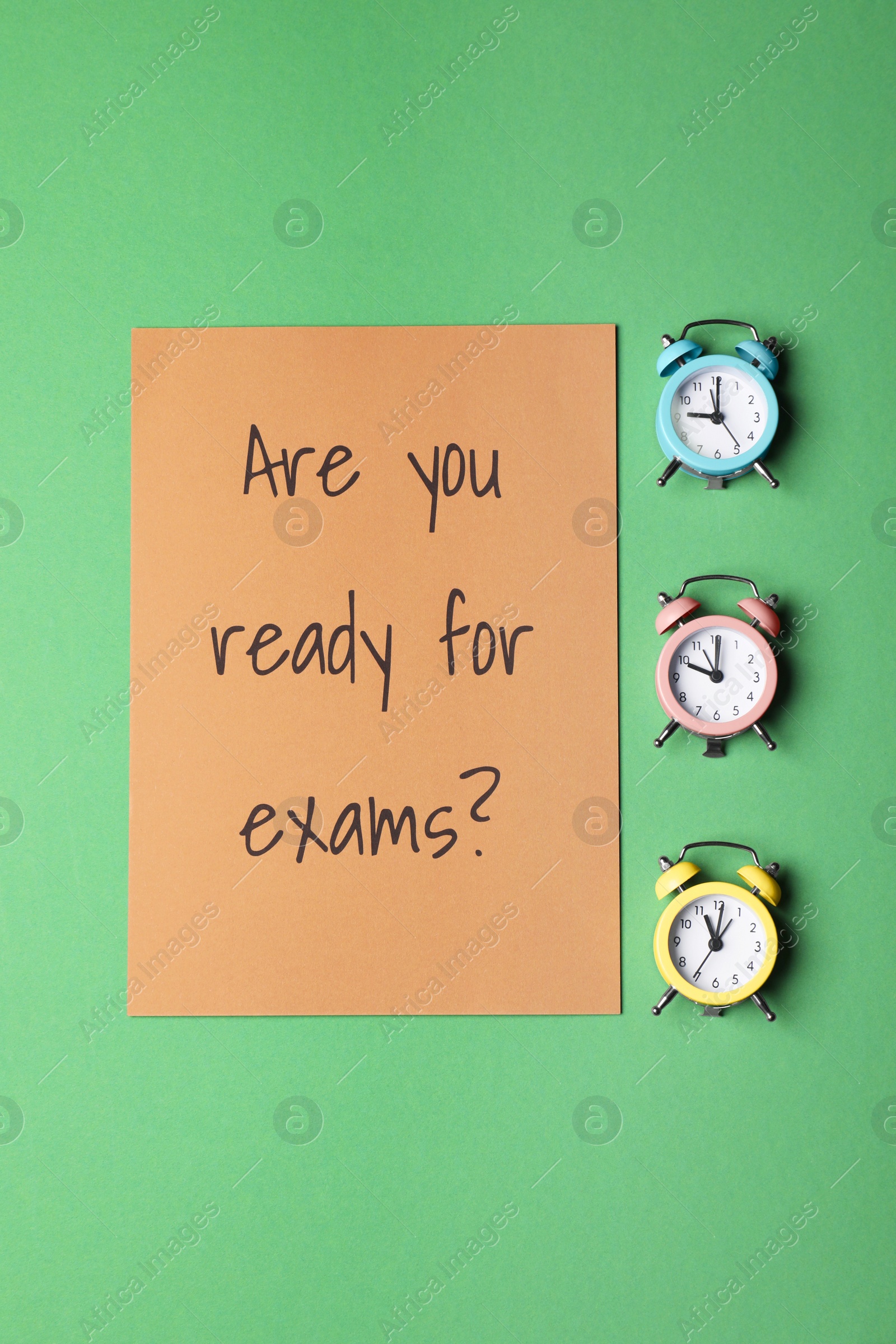 Photo of Paper with question Are you ready for exams and alarm clocks on green background, flat lay