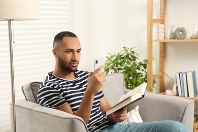 Photo of Young man using smartphone while reading book at home. Internet addiction