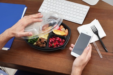 Office employee with smartphone having lunch at workplace, closeup. Food delivery