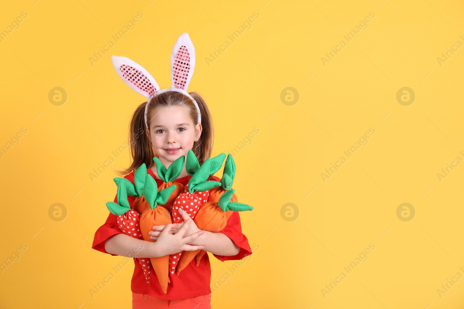 Photo of Adorable little girl with bunny ears and handful of toy carrots on orange background, space for text. Easter celebration