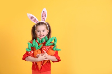 Photo of Adorable little girl with bunny ears and handful of toy carrots on orange background, space for text. Easter celebration