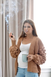 Photo of Young pregnant woman with cup of coffee smoking cigarette at home. Harm to unborn baby