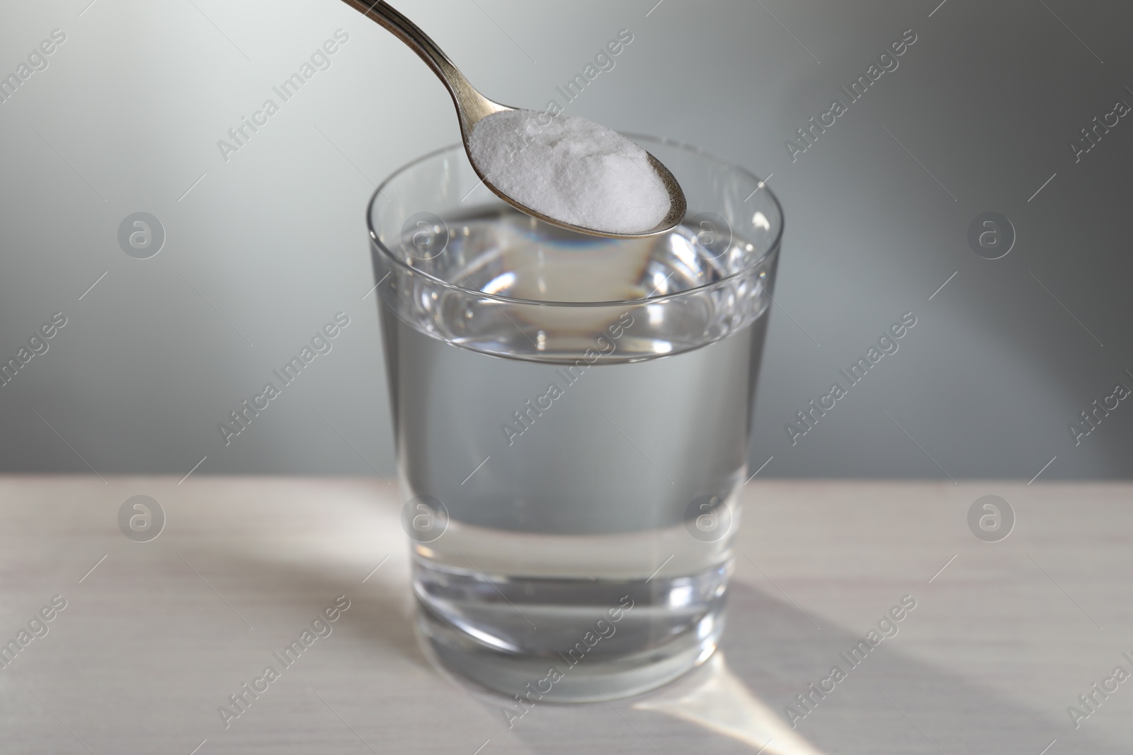 Photo of Spoon with baking soda over glass of water on table against light grey background, closeup