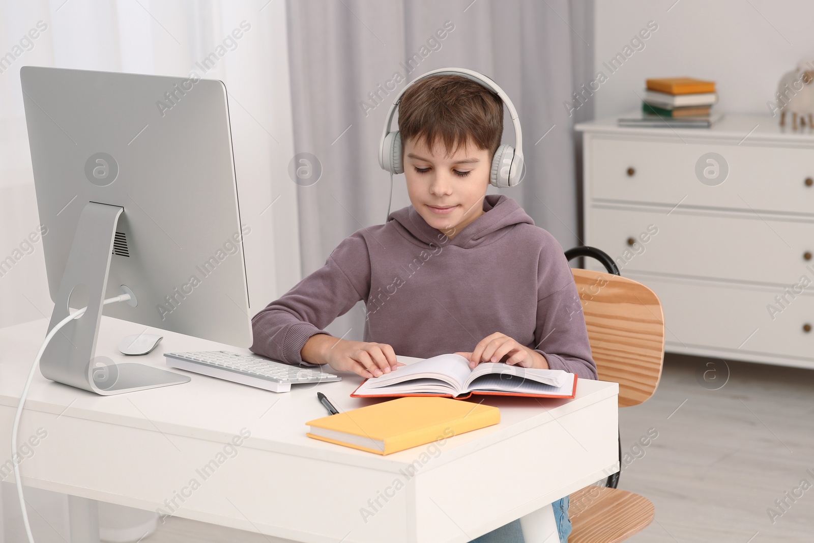 Photo of Boy reading book near computer at desk in room. Home workplace