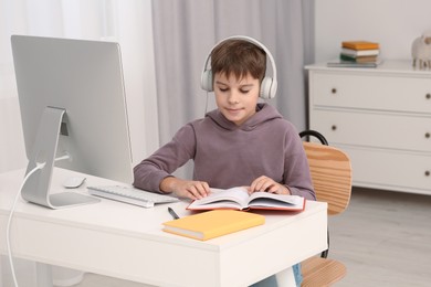 Boy reading book near computer at desk in room. Home workplace