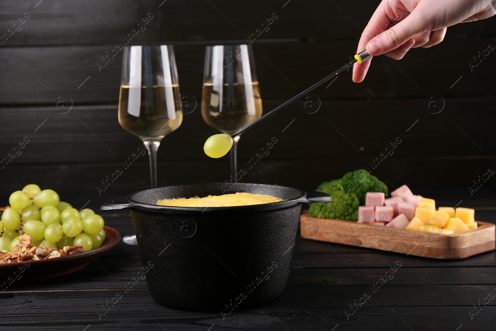 Photo of Woman dipping grape into fondue pot with melted cheese at black wooden table, closeup