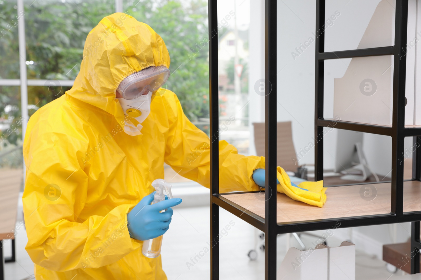 Photo of Janitor in protective suit disinfecting office furniture to prevent spreading of COVID-19